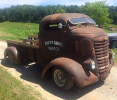 an old rusted truck parked on the side of a dirt road next to a field