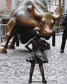 a woman standing next to a statue of a bull on a brick sidewalk with a man in the background