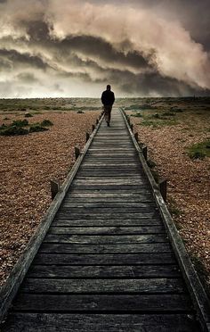 a man walking down a long wooden walkway under a dark sky with storm clouds in the background