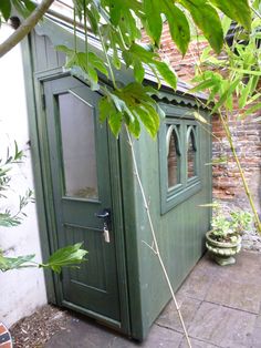 a small green outhouse sitting next to a tree and brick wall with potted plants