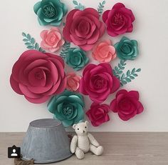 a teddy bear sitting next to a vase with paper flowers on the wall behind it