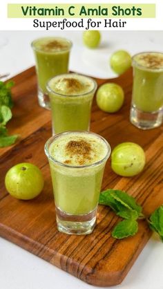 three glasses filled with green smoothie on top of a wooden cutting board next to fruit