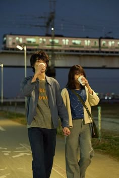 a man and woman walking down the street while holding coffee cups in their hands, with a train passing by