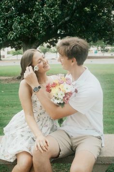 a young man and woman sitting on a bench in front of a tree with flowers