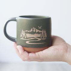 a hand holding a green coffee mug with mountains and trees on it