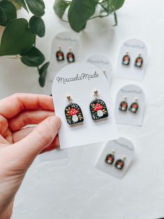 a person holding up some small earrings on top of a white table next to plants