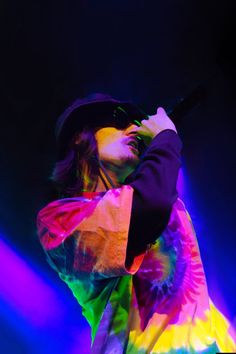 a man holding a microphone in his right hand and wearing a tie dye outfit on stage