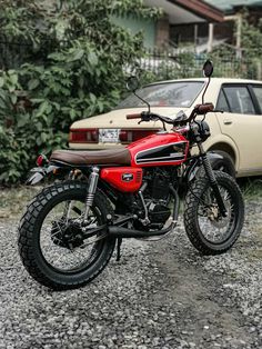 a red motorcycle parked in front of a car on gravel road next to shrubbery