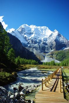 a wooden bridge over a river in front of a mountain with snow on it's top