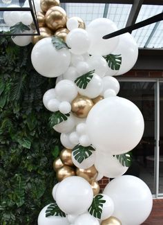 white and gold balloons are hanging from the ceiling in front of a green plant wall