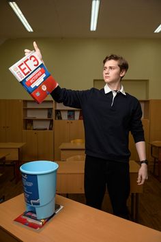 a man is holding up a book in front of a trash can on a table