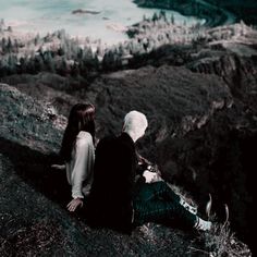 two people sitting on top of a hill looking at the water