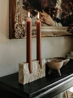 two candles sitting on top of a table next to a bowl and framed painting in the background