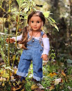 a doll is standing in the grass near a plant with long brown hair and blue overalls