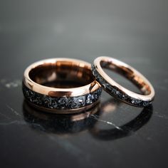 two gold wedding bands with black diamonds on them sitting on a marble countertop in front of a dark background