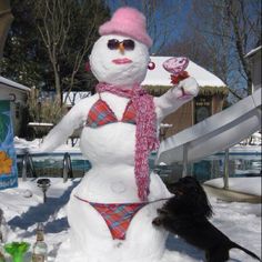 a snowman with a pink hat and scarf standing next to a dog in the snow