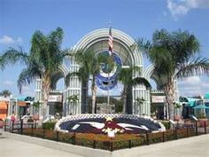 the entrance to an amusement park with lots of palm trees and flowers in front of it