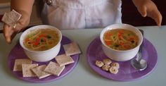 the doll is sitting at the table with two bowls of soup and crackers in front of her