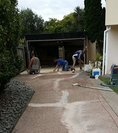 three men are working on the driveway in front of a house with trees and bushes