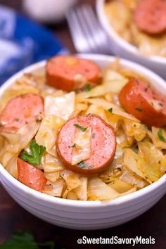 two white bowls filled with sausage and cabbage soup on top of a wooden table next to silverware