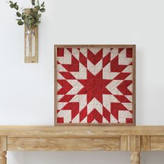 a red and white quilt on a wooden table with a vase full of flowers in the background