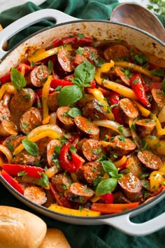 a pan filled with sausage and peppers on top of a green cloth next to bread