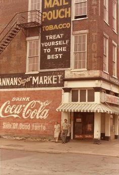 an old photo of two men standing in front of a coca - cola advertisement on the side of a building