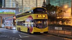 a yellow double decker bus driving down a street next to tall buildings at night time