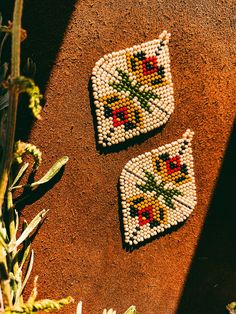 two pieces of beaded art sitting on top of a dirt ground next to plants