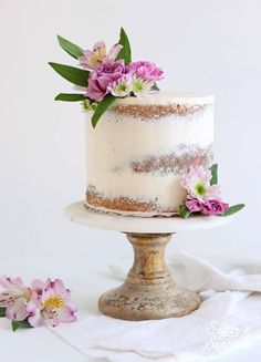 a white cake with pink flowers on top sitting on a wooden stand next to a napkin