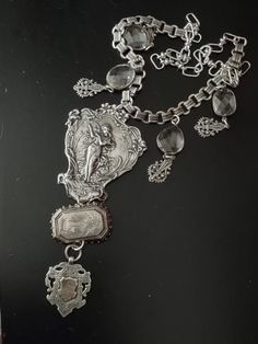 The central pendant in this assemblage is an art nouveau plaque in plated silver that most likely adorned the back of a mirror. It supports an antique Victorian aesthetic movement sterling brooch featuring a flying swallow. From the brooch is a hallmarked British sterling Victorian era watch fob with a gilded initialed cartouche. Accompanying pendants are rhodium plated bezel set free form faceted crystals and Victorian inspired filigree charms. All elements are attached to silver plated book chain and the necklace is finished with larger links and a hook so you can adjust the length. Elegant Silver Medallion Brooch, Silver Art Deco Medallion Necklace, Victorian Silver Necklace With Antique Finish, Silver Medallion Art Deco Jewelry, Victorian Silver Necklace For Collectible, Victorian Silver Necklace, Art Deco Silver Jewelry With Vintage Charm, Silver Art Deco Jewelry With Vintage Charm, Antique Silver Pendant Brooches