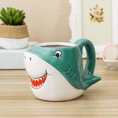 a ceramic mug with a shark face painted on the front and side, sitting on a wooden table