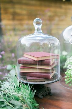 several books are under a glass clochel on a table with flowers and greenery