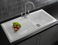 a white kitchen sink with two faucets and some fruit on the counter top