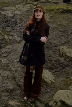 a woman standing on top of a rocky hillside wearing a hat and coat with her arms crossed