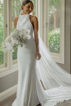 a woman in a white wedding dress standing by a window with her veil over her shoulder