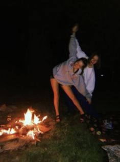 two people standing next to a campfire at night with their arms in the air