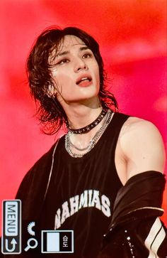 a young man with long hair wearing a black shirt and chain around his neck, standing in front of a red background