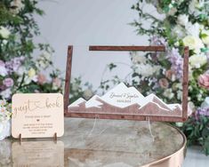 a wooden frame with mountains on it sitting next to a card holder and flower arrangement