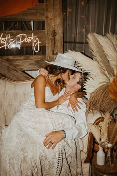 two women in white dresses and hats kissing each other on a couch with feathers hanging from the ceiling