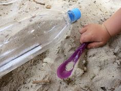 a baby reaching for a purple spoon in the sand next to a plastic water bottle