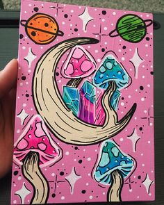 a hand holding up a small painting on a pink background with mushrooms, planets and stars