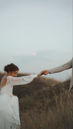 a bride and groom holding hands in a field with tall grass on the other side