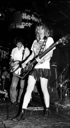 a woman playing guitar while standing on stage with other people behind her in black and white