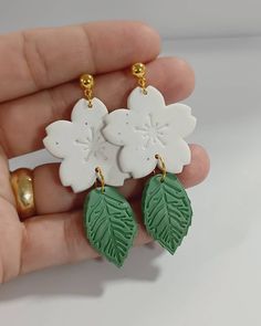 a pair of white and green flower earrings with leaves on each earring, held in someone's hand