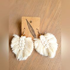 two white tasseled earrings on top of a wooden table next to a tag