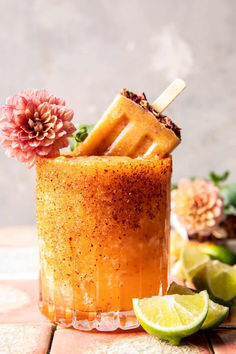 an orange cocktail garnished with a flower and two lime wedges on the side