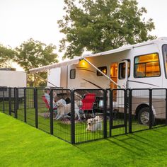 an rv is parked behind a fence with chairs and a dog on the grass in front of it