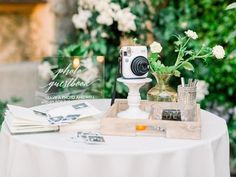 a white table topped with a vase filled with flowers next to a camera and other items