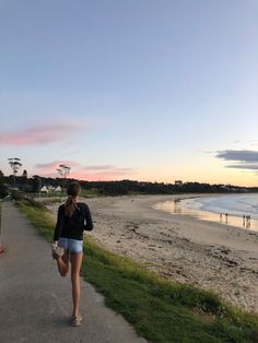 a woman walking down a sidewalk next to the ocean at sunset or sunrise with people swimming in the water